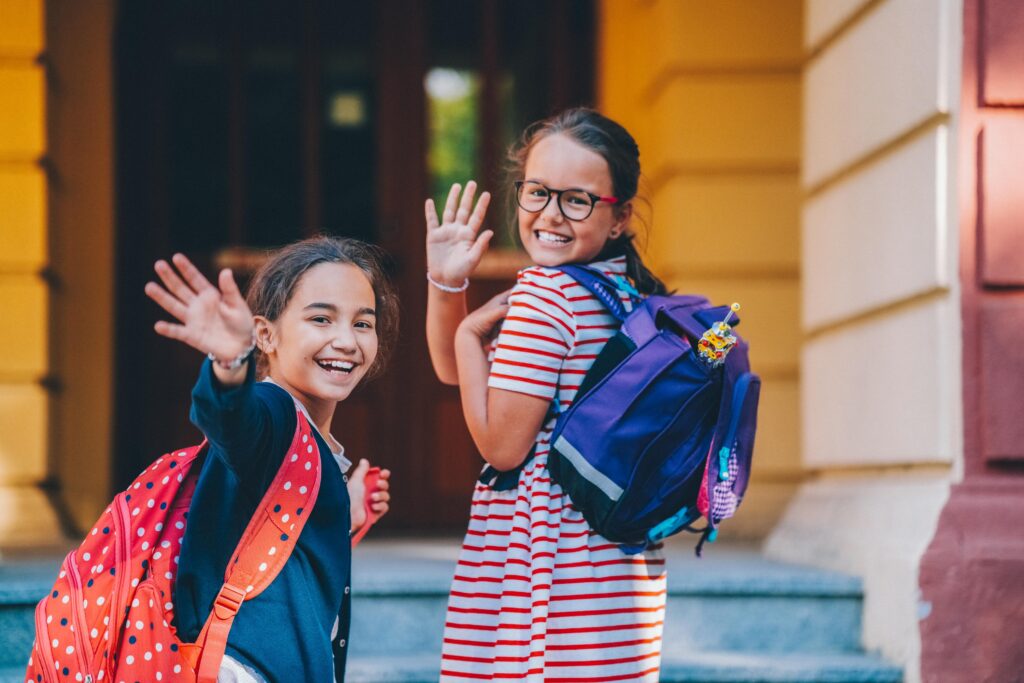 two girls waving goodbye walking into school